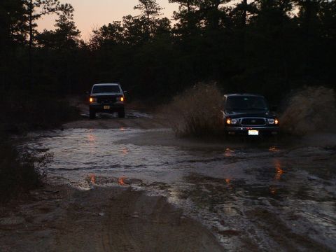 1994 Ford Bronco - Two SUVs splash through a water obstacle on a dirt road as the sun sets.