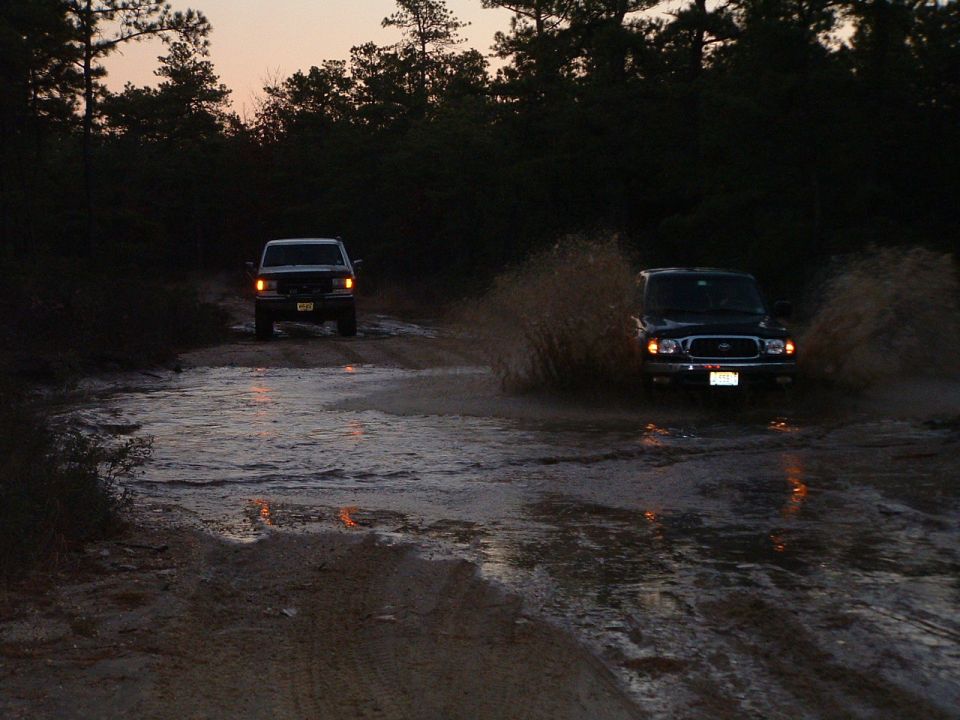 two SUVs driving through a muddy water crossing at dusk