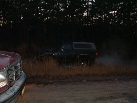 1994 Ford Bronco - A dark SUV with a camper shell is seen from the side, kicking up dust as it drives. Another vehicle is partially visible in the foreground.