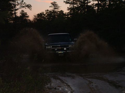 1994 Ford Bronco - A dark SUV drives through a muddy path, kicking up water and dirt as the sun sets.