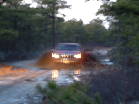 1994 Ford Bronco - A red SUV navigates a muddy trail, kicking up water and dirt. The vehicle's headlights illuminate the scene.