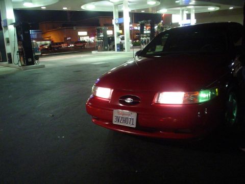1993 Ford Taurus - A red 1993 Ford Taurus is parked at a gas station at night, with its headlights on.