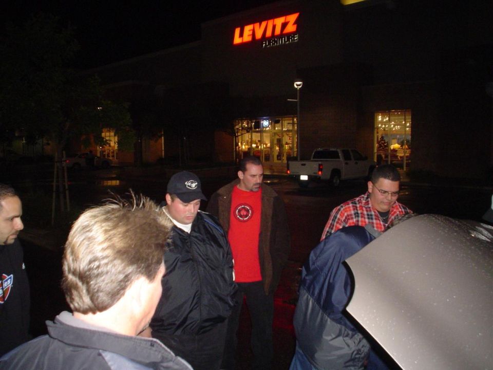 Guys looking at car hood open outside Levitz Furniture store at night. White pickup truck parked nearby