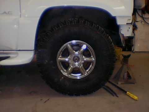 1999 Cadillac Escalade - Close-up of a white SUV's front wheel and part of the bodywork, with a jack stand visible.