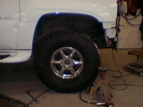 1999 Cadillac Escalade - A white SUV's front wheel and fender are visible, with the front bumper removed and the vehicle supported by a jack stand.