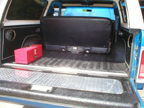 1978 Ford Bronco - Rear cargo bay of a blue SUV, tailgate open, with a red metal toolbox, a black fold-down seat and a diamond-plate sill.