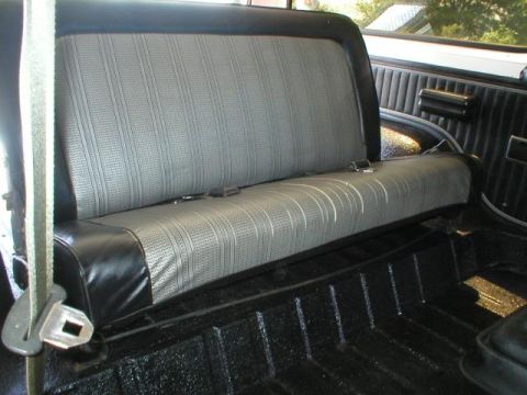 1978 Ford Bronco - Interior rear bench seat with gray ribbed cloth upholstery and black vinyl trim, showing a lap seat belt with metal buckle and a bare black…