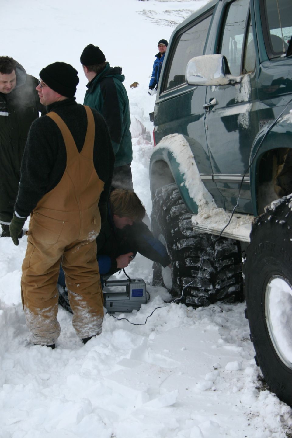 Dark green Bronco with big off-road tires stuck in deep snow as men in winter gear work on it