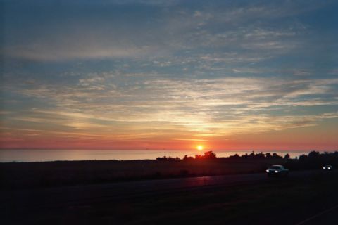 1990 Ford Bronco - vivid orange sun sets over a flat hazy horizon under streaked clouds, a single car visible on the road in the foreground.