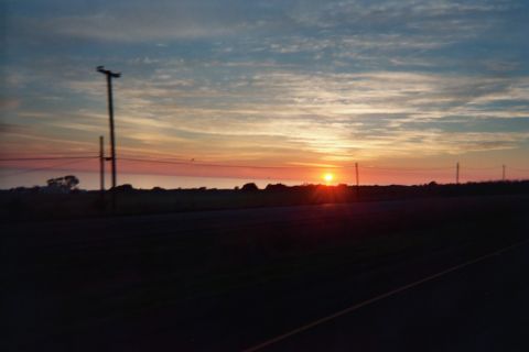 1990 Ford Bronco - sun sets over flat farmland, a utility pole and power lines silhouetted against orange and blue streaked clouds beside a roadway.