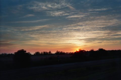 1990 Ford Bronco - sun sets behind low trees over flat fields, casting orange and blue across streaked clouds above foreground railroad tracks.