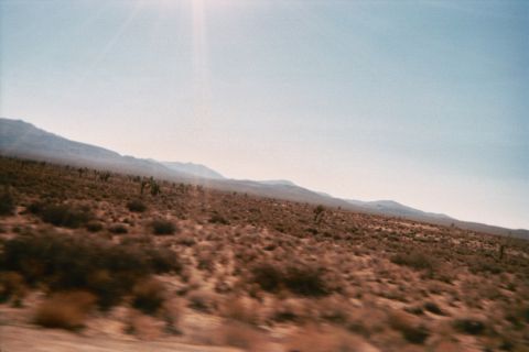 1990 Ford Bronco - Sunlit desert scrub with sparse brush and distant mountains under a hazy blue sky, shot from a moving vehicle.