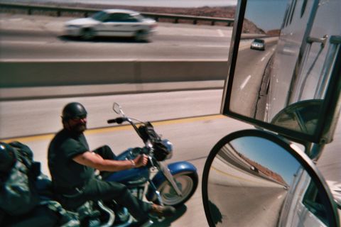 1990 Ford Bronco - helmeted rider on a blue cruiser motorcycle travels a multi-lane highway; a car passes and a side mirror frames the right edge.