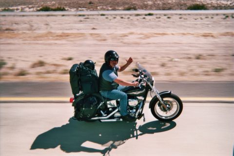 1990 Ford Bronco - rider in jeans and helmet waves from a black cruiser motorcycle loaded with gear, speeding along a desert road.