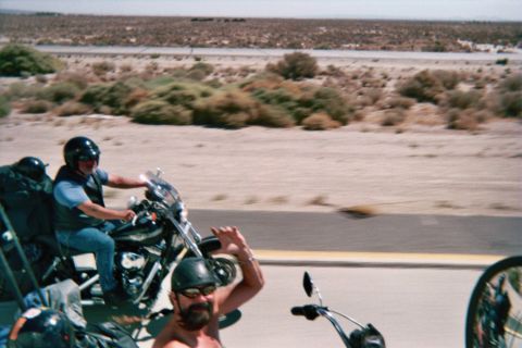 1990 Ford Bronco - Two helmeted riders on black motorcycles travel a desert highway, one foreground rider raising a hand near a chrome handlebar.