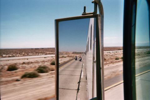1990 Ford Bronco - Side mirror reflection showing two motorcycles trailing on a straight desert highway with distant mountains.