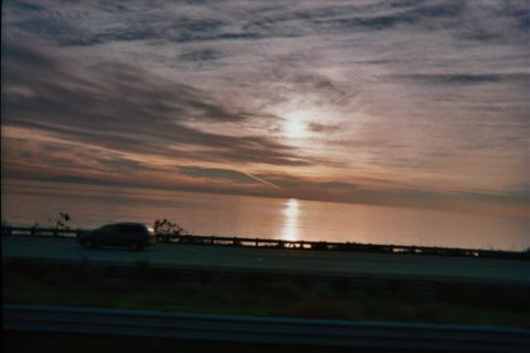 1990 Ford Bronco - cloudy orange sunset reflects on water beside a guardrail-lined highway as a silver car drives past in low light.