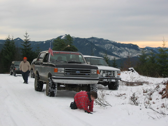 Lifted two-tone Ford Bronco carrying a tree on a snowy mountain trail with people nearby