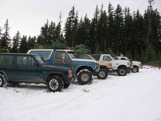 Row of lifted 4x4 SUVs in snow by pine forest, Jeep Cherokee in front