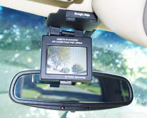 Aftermarket backup camera monitor mounted above the rear-view mirror inside a vehicle, screen showing trees behind.