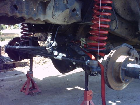 1995 Ford Bronco - Under-truck view of a coil-sprung solid front axle supported on red jack stands, with the front hub, rotor and wheel studs exposed on the r…