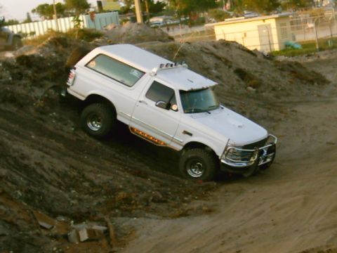 1995 Ford Bronco - High-angle passenger-side three-quarter view of a white lifted Ford Bronco descending a dirt mound in a sandy lot. Visible are a chrome tub…