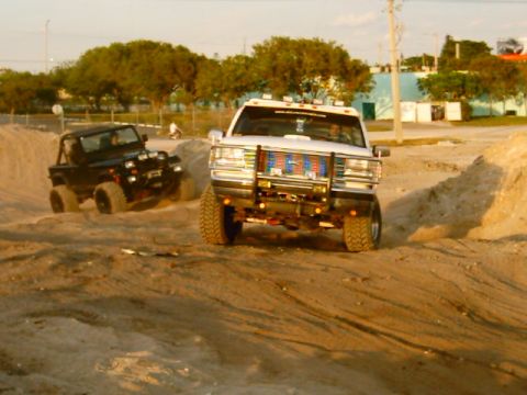 1995 Ford Bronco - White lifted Ford Bronco climbing out of a sand pit head-on, wearing a chrome brush guard with an American-flag grille insert, roof-mounted…