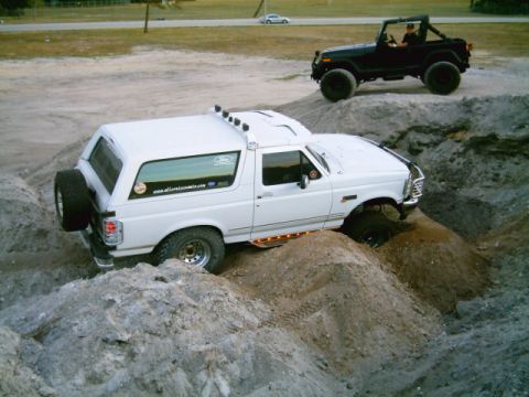 1995 Ford Bronco - Elevated view of a white lifted Ford Bronco flexing over a dirt mound course, front wheel dropped into a hollow with the rear tire lifting.…