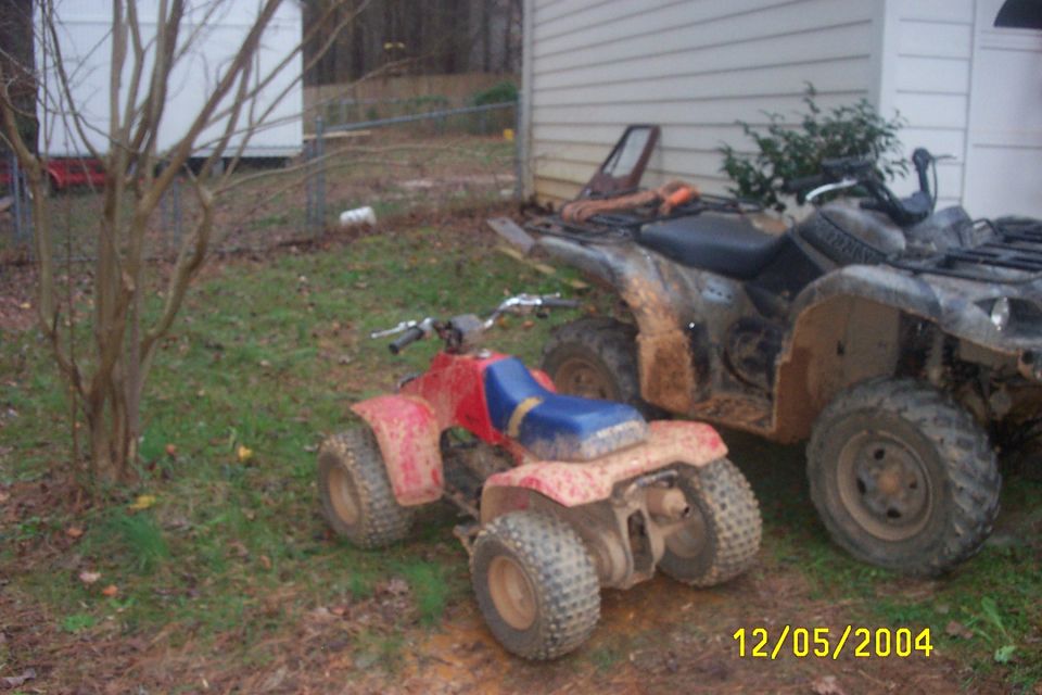 Two muddy ATVs parked in a grassy yard next to a building. A smaller red and blue ATV is in the foreground, and a larger dark ATV is behind it