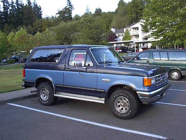 Blue and black Ford Bronco parked in a lot. 'For Sale' sign in window