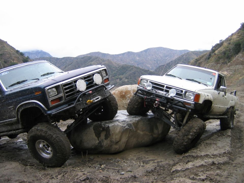 Blue Ford Bronco and white Toyota pickup on lifted tires crawling over a boulder on a muddy mountain trail