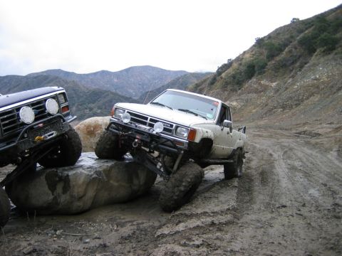 1985 Ford Bronco - Two modified trucks navigate a challenging off-road trail on a muddy hillside.