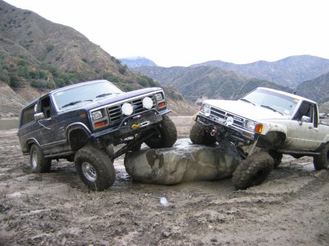 1985 Ford Bronco - Two modified off-road vehicles navigate a challenging rocky and muddy terrain in a mountainous area.