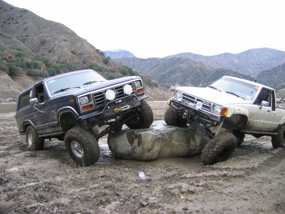 Blue Ford Bronco and white Toyota pickup crawling over a boulder in muddy off-road terrain
