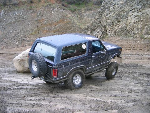 1985 Ford Bronco - A dark blue SUV navigates a muddy, off-road environment with a spare tire mounted on its rear.