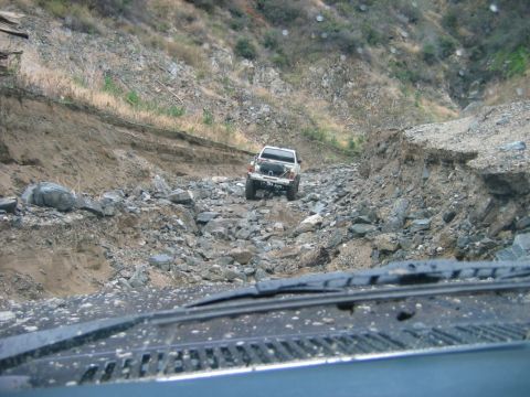 1985 Ford Bronco - A white pickup truck navigates a treacherous, rocky mountain pass, with another vehicle's dashboard visible in the foreground.