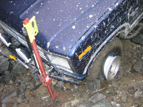1985 Ford Bronco - A blue off-road vehicle is stuck on a rocky terrain, with a jack positioned on its hood and a flat tire.
