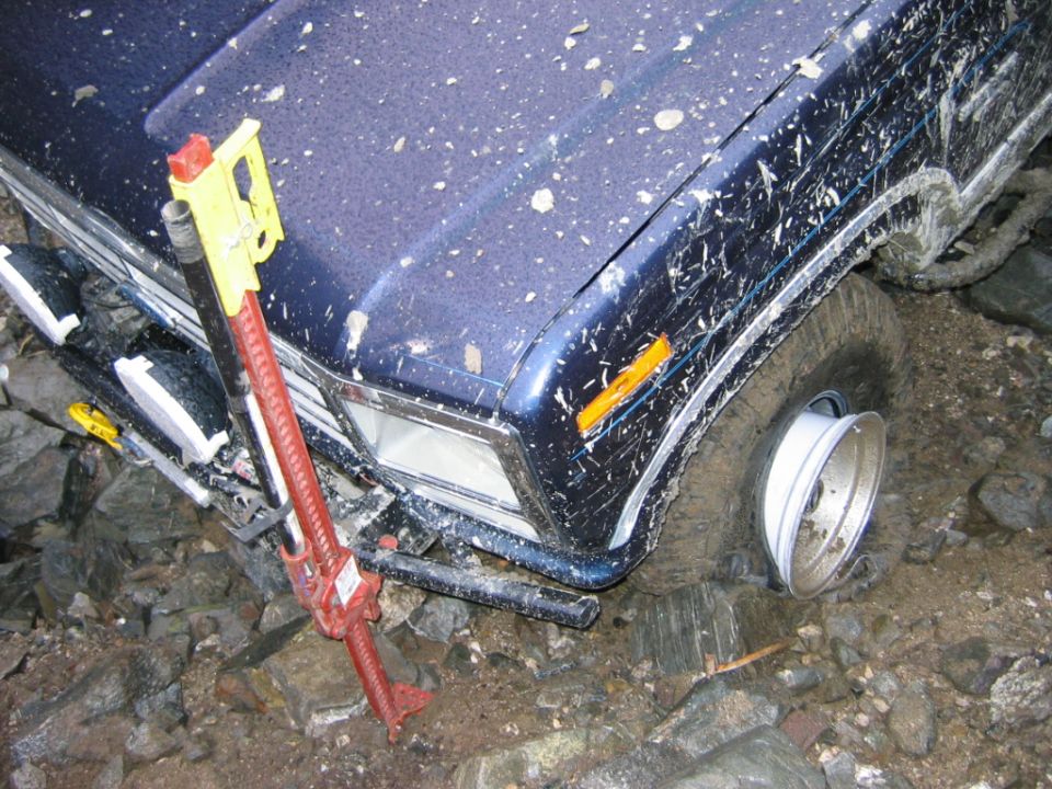 Blue Bronco front on rocks with red high-lift jack and bare steel wheel on ground