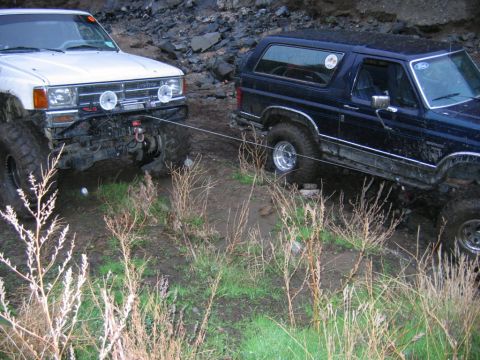 1985 Ford Bronco - A dark blue Ford Bronco is stuck in the mud, with a white Toyota truck attempting to pull it out.