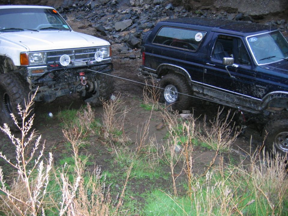 Dark blue Ford Bronco winch-cabled to a white Toyota pickup on muddy off-road terrain