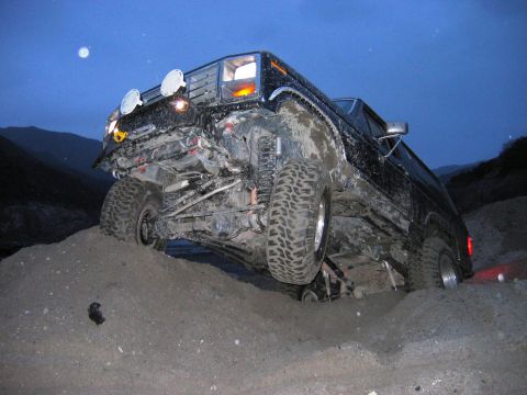 1985 Ford Bronco - Off-road SUV with significant front-end damage stuck in loose dirt under a dark sky.