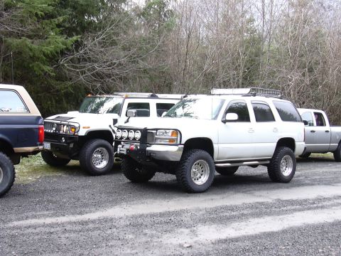 1991 Ford Bronco - Multiple off-road vehicles are parked together on a gravel surface, with a wooded area behind them.