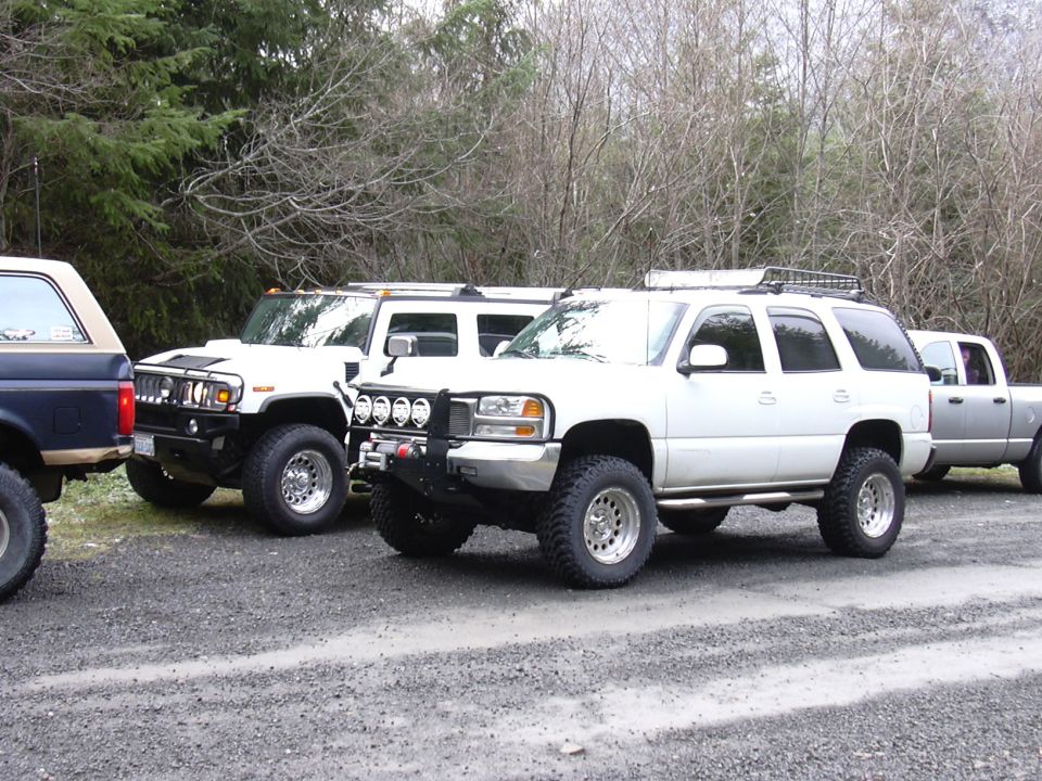 Several modified SUVs and a pickup truck parked on a gravel lot with trees in the background