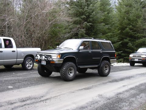 1991 Ford Bronco - A dark-colored SUV with off-road tires drives on a gravel road, accompanied by other vehicles in a wooded area.