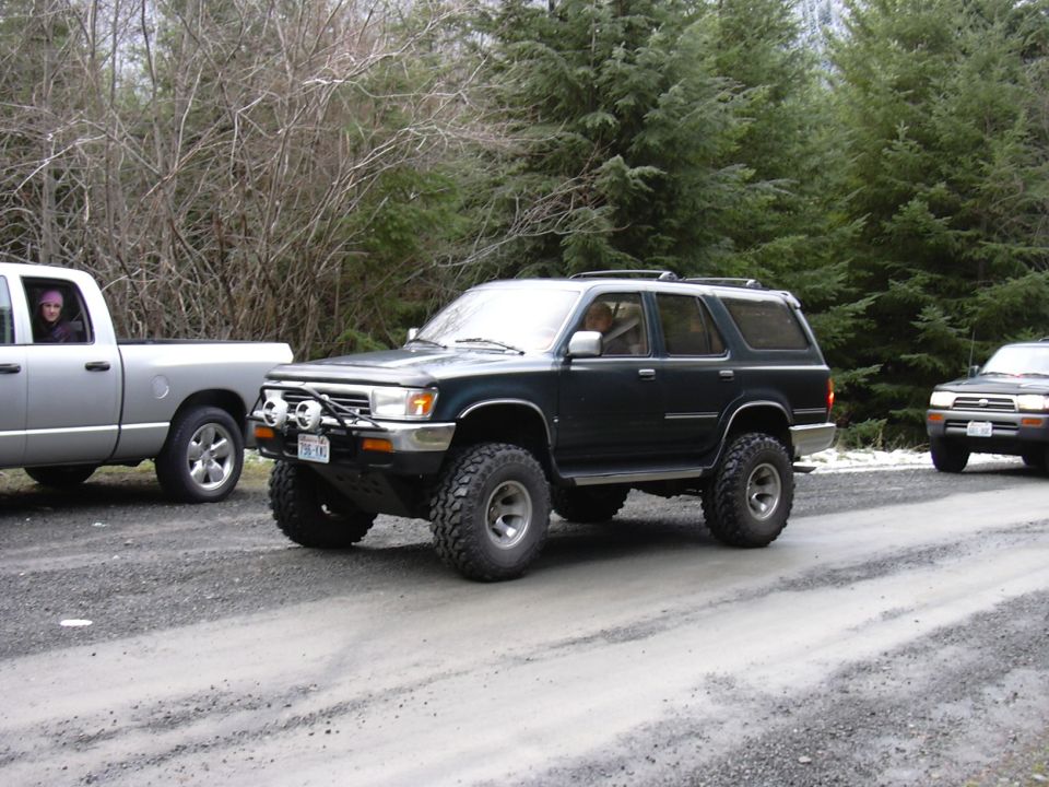 Dark SUV with big tires on a dirt road, silver pickup truck to its left, another SUV behind it