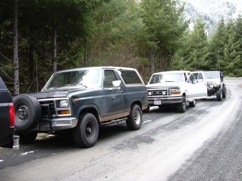 1991 Ford Bronco - A line of vehicles, including a green Ford Bronco, are parked on a dirt road next to a forest.