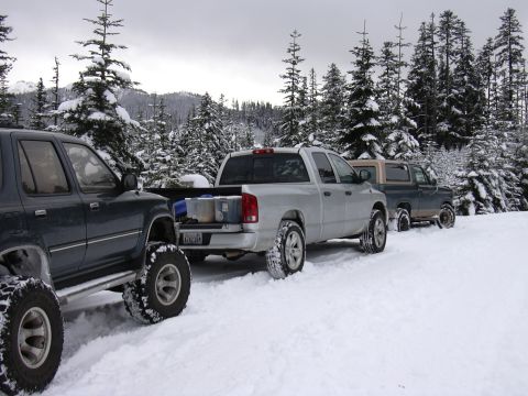 1991 Ford Bronco - A line of off-road vehicles are parked on a snow-covered road surrounded by pine trees.