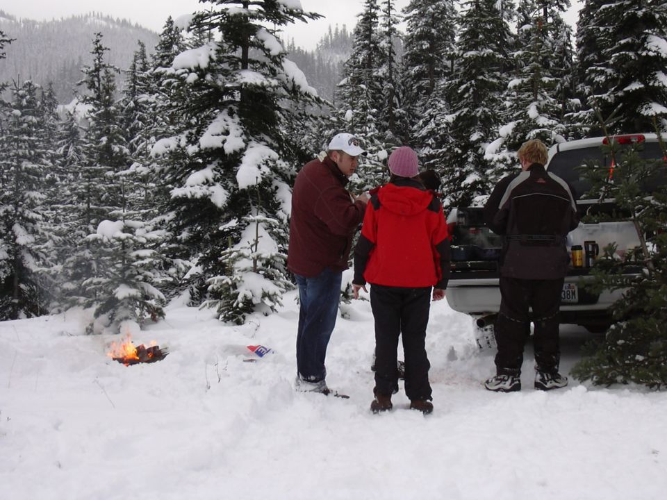 three people stand by a pickup truck with a campfire going in a snowy forest