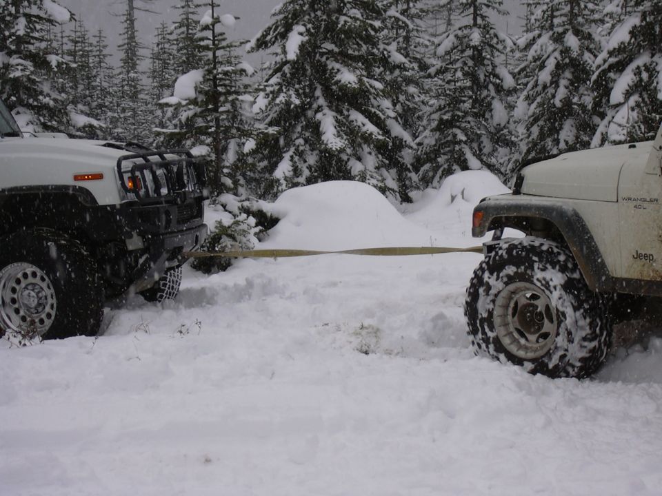 White Jeep Wrangler and another white SUV being towed by a strap in a snowy forest