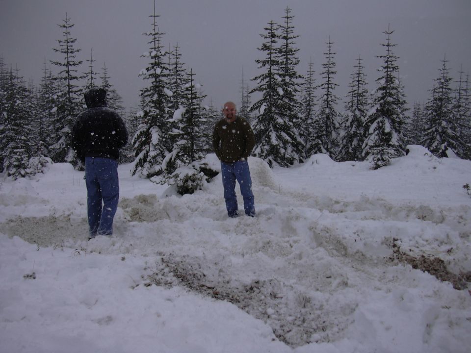 Two people stand in a snowy forest with snow-covered trees and ground
